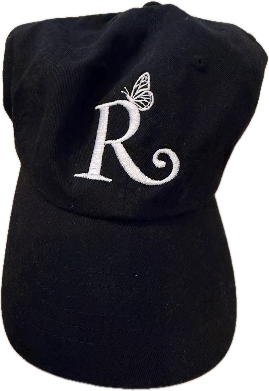 Black baseball cap with the white embroidered Rebellious Aging R logo, a butterfly resting on top of the letter.