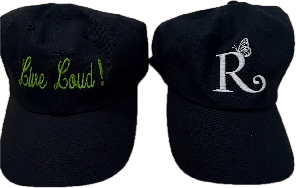 The two Rebellious Aging hats side by side: a black cap with “Live Loud!” in green script, and a black cap with the white R and butterfly logo.